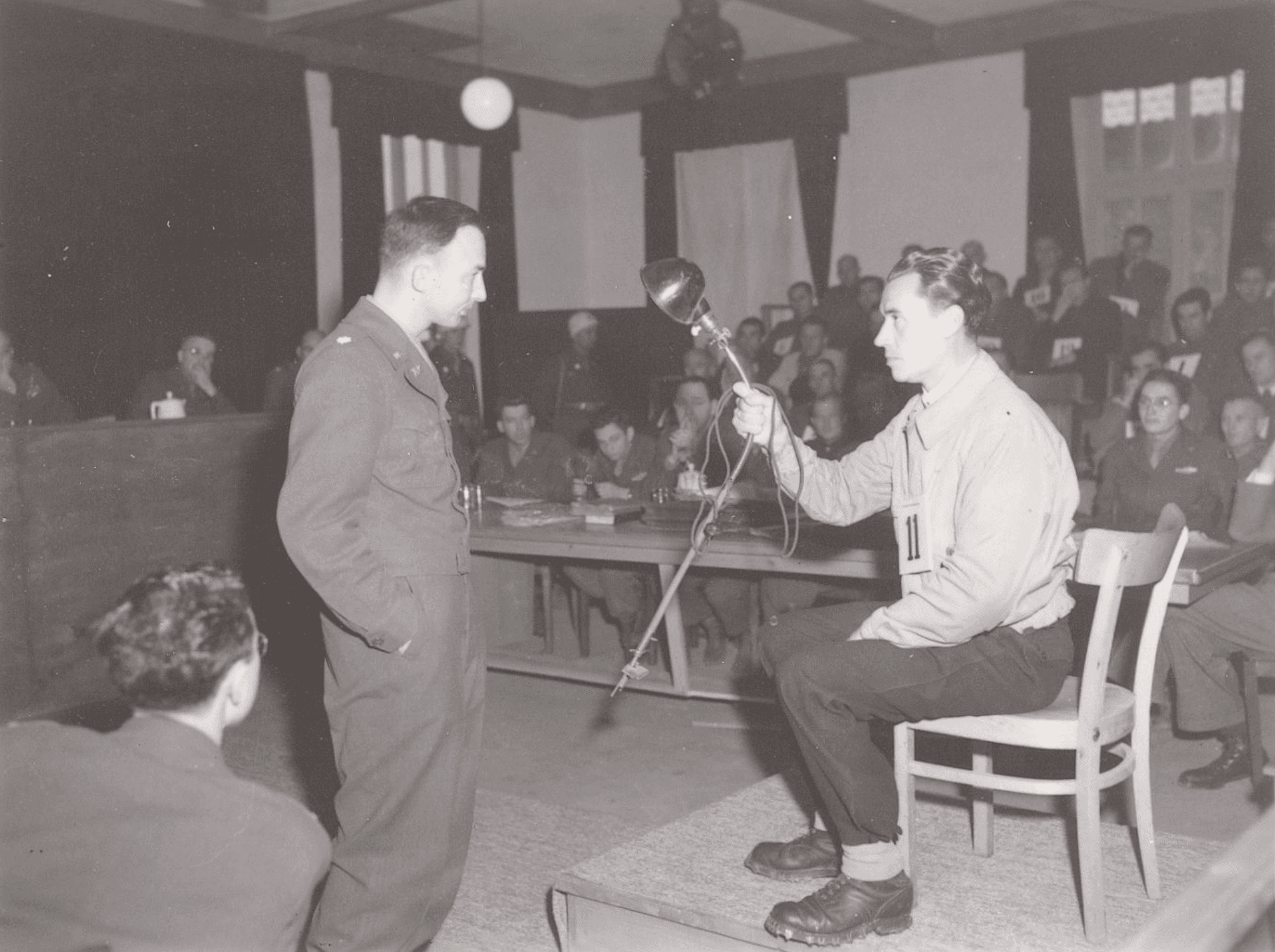 Black and white photo of courtroom scene. One man is seated on a platform holding a metallic object.